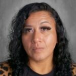 A close-up portrait of a woman with curly black hair, wearing a dark shirt and patterned cardigan, against a gray background.