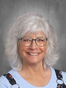 Smiling woman with white hair and glasses, wearing a light blue top and patterned overalls, against a gray background.