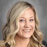Smiling woman with long, wavy blonde hair, wearing a black top, against a gray background.