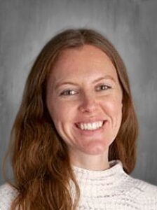 Headshot of a smiling woman with long wavy hair, wearing a white sweater, against a gray background.