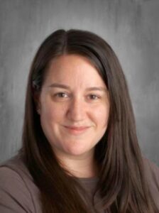 Portrait of a woman with long brown hair, wearing a gray shirt, smiling against a neutral background.