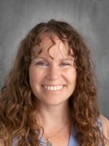Smiling woman with curly brown hair, wearing a light blue top, against a gray background.