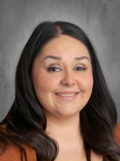 A smiling woman with long dark hair wearing a brown jacket, set against a gray background.