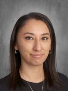 A woman with long dark hair and earrings smiles softly against a gray background. She wears a black shirt.