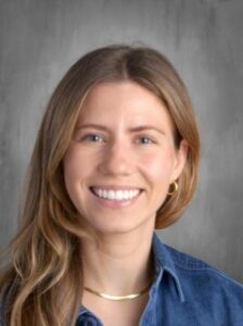 Smiling woman with long, wavy hair, wearing a denim shirt and gold hoop earrings against a gray background.
