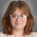 A young person with medium-length, wavy hair and glasses, smiling against a gray background.