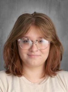 Una persona joven con el pelo medio largo y ondulado y gafas, sonriendo sobre un fondo gris.