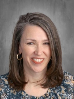 A smiling woman with medium-length, straight gray hair, wearing hoop earrings and a floral top, against a gray background.