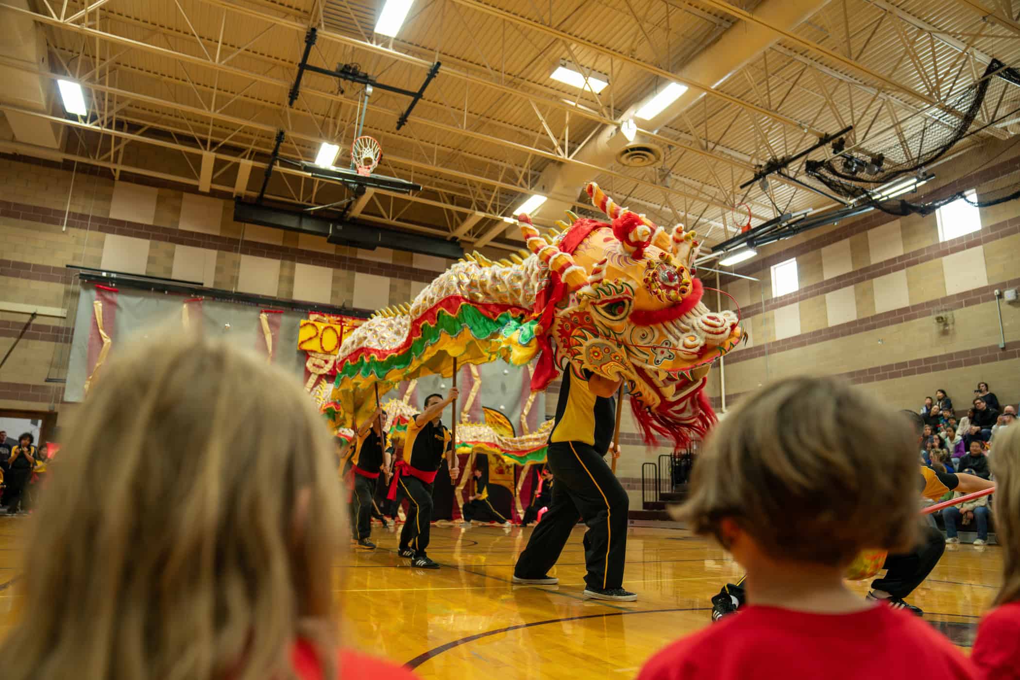 A colorful dragon puppet is paraded in a gym during a performance, with children watching in the foreground.