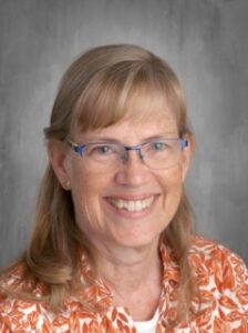 A smiling woman with shoulder-length light brown hair, wearing glasses and an orange patterned shirt, against a gray background.