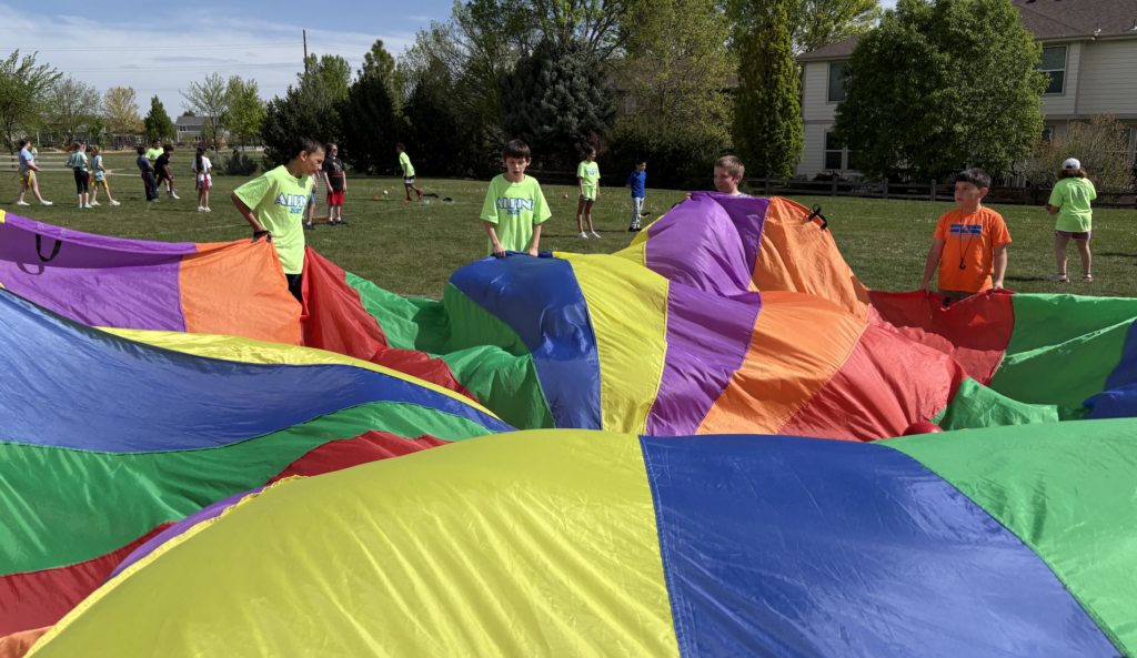 Multiple kids in bright shirts hold a large, colorful parachute on a grass field during an outdoor activity.