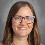 A woman with long brown hair and glasses smiles at the camera, wearing a light brown sweater.