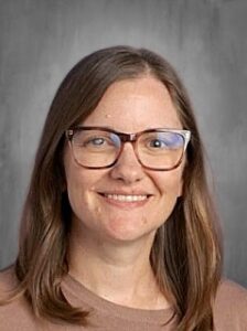 Una mujer de pelo largo castaño y gafas sonríe a la cámara, con un jersey marrón claro.