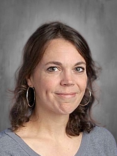 Headshot of a woman with brown hair, wearing hoop earrings and a gray shirt, smiling against a gray background.
