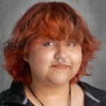 Young person with wavy, orange hair, a septum piercing, and wearing a black zip-up jacket, smiling at the camera.