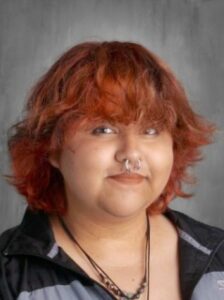 Young person with wavy, orange hair, a septum piercing, and wearing a black zip-up jacket, smiling at the camera.