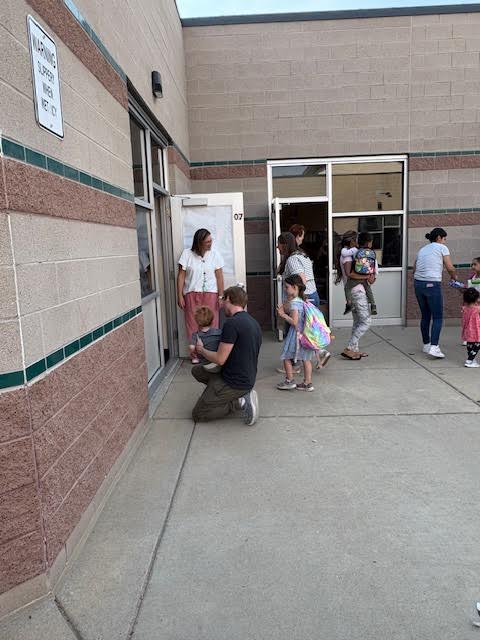 A group of children and adults gather outside a school entry, with one adult kneeling next to a child.
