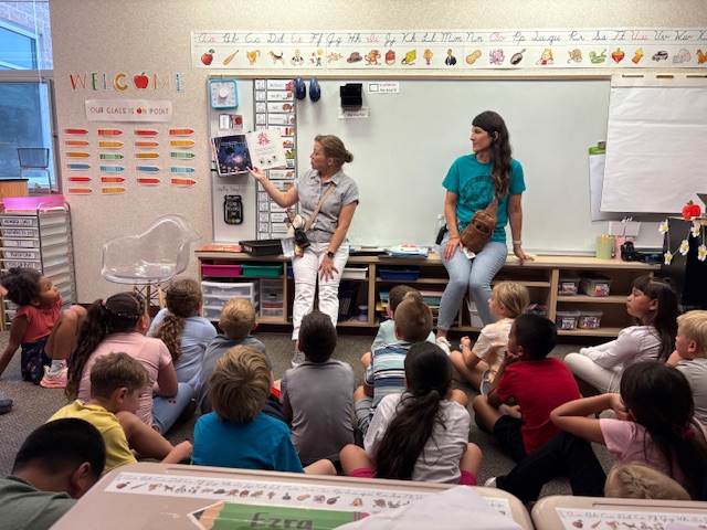 Two women present to a group of seated children in a colorful classroom filled with educational materials.