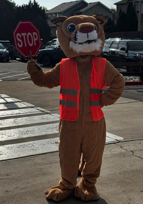 A person in a cartoon animal costume wears an orange vest and holds a stop sign, standing at a crosswalk.