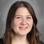A smiling young woman with long brown hair, wearing a dark sweater, against a neutral background.