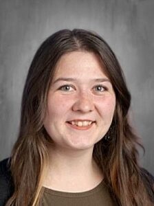 A smiling young woman with long brown hair, wearing a dark sweater, against a neutral background.