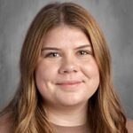 A young woman with long, wavy brown hair smiles softly at the camera, set against a gray background.