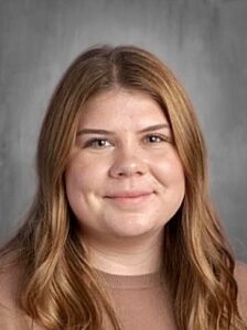 A young woman with long, wavy brown hair smiles softly at the camera, set against a gray background.