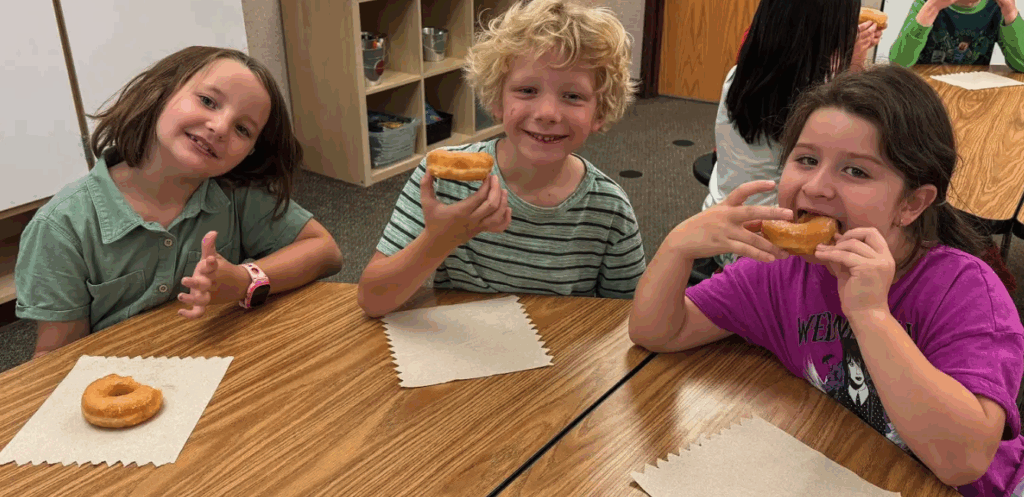 Three children smile while enjoying donuts at a table, with one girl playfully posing and the others holding their treats.