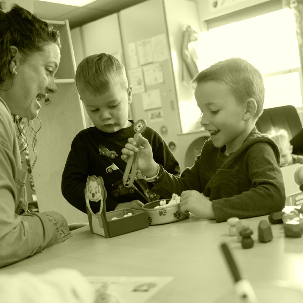 A woman engages with two young boys at a table, where they play with toys and craft materials in a bright classroom.