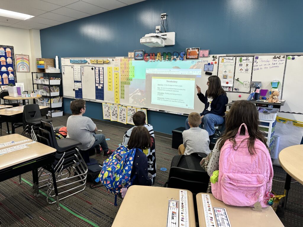 A teacher instructs a group of students sitting on the floor in a colorful classroom, with a projector displaying vocabulary.