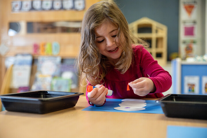A young girl with long hair smiles while gluing colorful paper pieces onto a blue sheet at a classroom table.