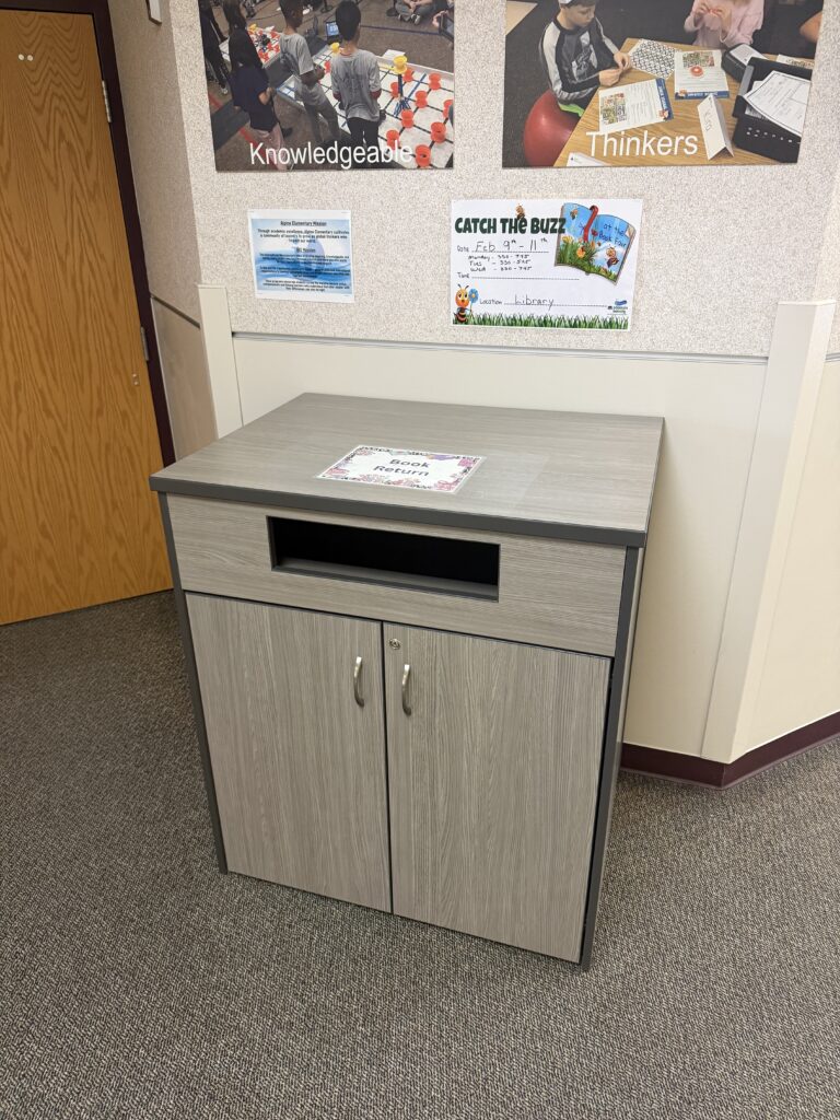 A gray book return cabinet with a top slot and two doors, located in a library setting with educational posters nearby.