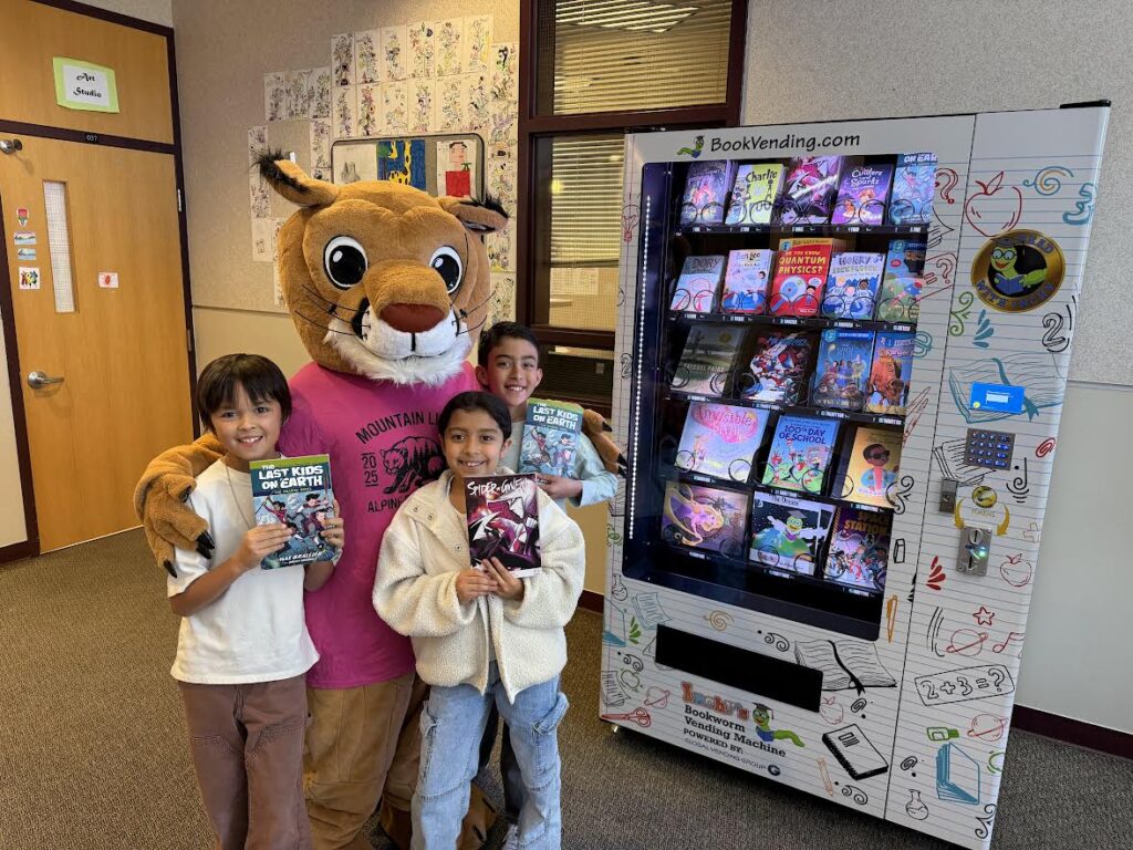 Children pose with a mascot next to a colorful book vending machine displaying various titles.