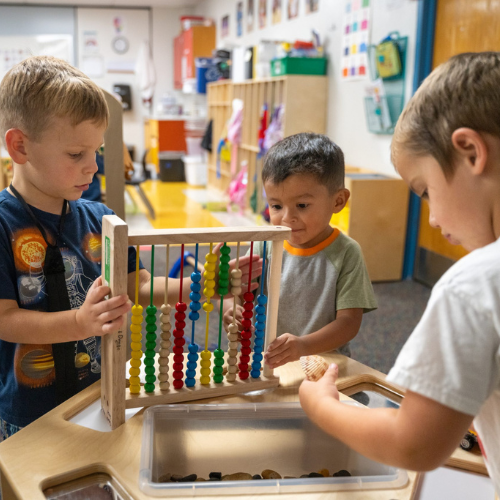 Tres niños pequeños juegan con un ábaco de madera y una caja de juguetes en un colorido aula.