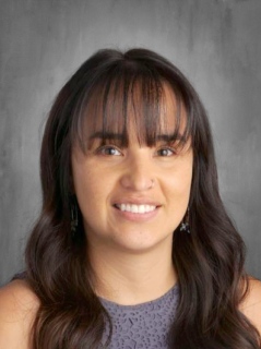 A smiling woman with long dark hair and bangs, wearing a gray top, against a neutral background.