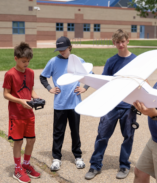 Three boys gather outdoors, one holding a remote control, while another prepares to launch a model airplane.